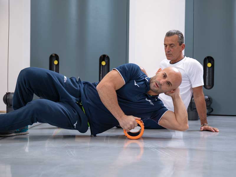 Personal trainer Show Club mostra un esercizio con foam roller sulla zona costale laterale del torace, mentre un uomo osserva il movimento.
