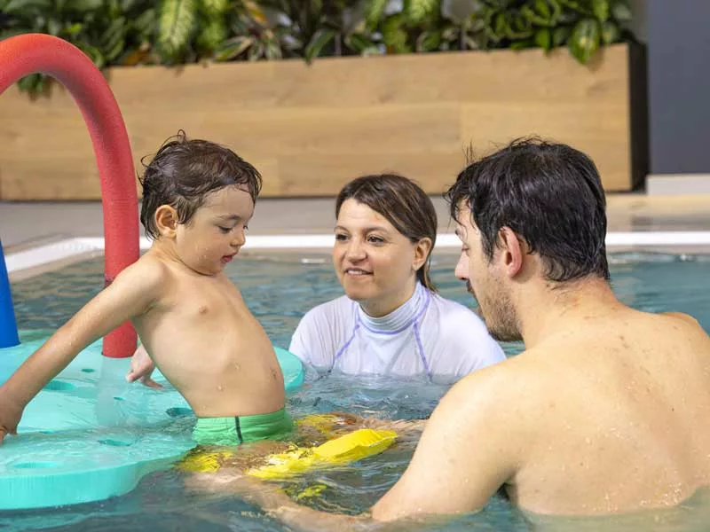 bambino di tre anni entra in piscina con il papà. Usa un salvagente perché non Sto arrivando!nuotare. C'è una personal trainer al loro fianco che li segue.  