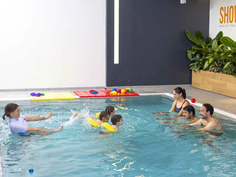 tre bambini di 2 e 3 anni fanno le loro prime nuotate in piscina usando il salvagente. I loro genitori sono in vasca che li aiutano nei loro primi passi in acqua. Tutto è sotto la supervisione della personal trainer 