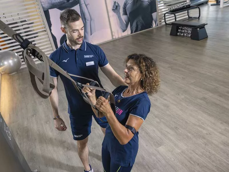 donna sorridente di 70 anni si allena in palestra a Padova usando il trx ed è seguita da un personal trainer che le consigli esercizi di ginnastica dolce