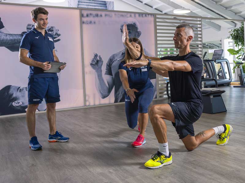 Uomo esegue una contro piegata in palestra, mantenendo l’equilibrio con il supporto di un personal trainer, durante una sessione di Training neuromotorio.