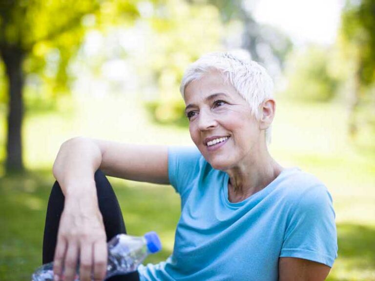 donna di 50 anni in menopausa è vestita sportiva e sta facendo una pausa seduta al parco. è sorridente.