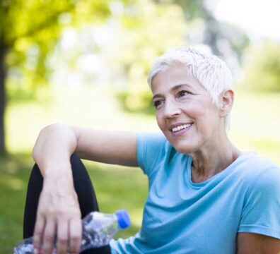 donna di 50 anni in menopausa è vestita sportiva e sta facendo una pausa seduta al parco. è sorridente.