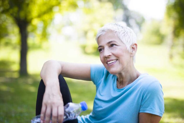 donna di 50 anni in menopausa è vestita sportiva e sta facendo una pausa seduta al parco. è sorridente.