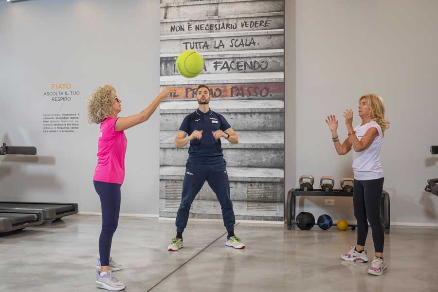 Due donne partecipano a un allenamento di gruppo cardio, lanciandosi la palla mentre il personal trainer le incoraggia.