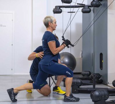 Una donna over 40 si allena in contro-piegamento con il Kinesis, lavorando su schiena e braccia, seguita da un personal trainer che controlla la postura.