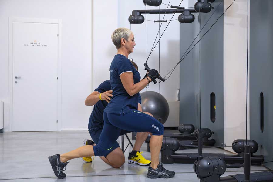 Una donna over 40 si allena in contro-piegamento con il Kinesis, lavorando su schiena e braccia, seguita da un personal trainer che controlla la postura.
