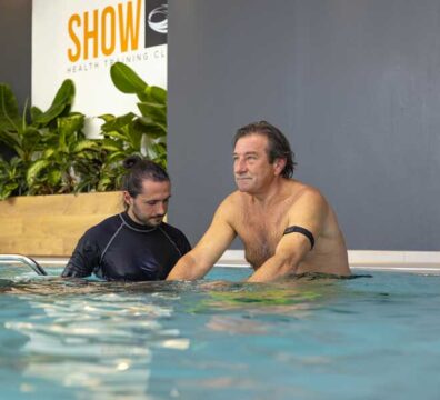 Un uomo esegue un allenamento posturale in acqua, seguito dal personal trainer che corregge la postura e guida l’esercizio.