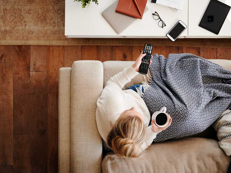 Donna sedentaria seduta sul divano con una coperta, tiene in mano il telecomando e una tazza di caffè mentre guarda la televisione.