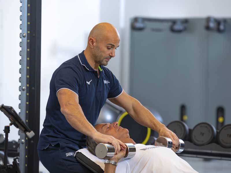 Uomo esegue un allenamento di forza per le braccia con i pesi su panca in palestra, assistito da un personal trainer durante l’esecuzione.