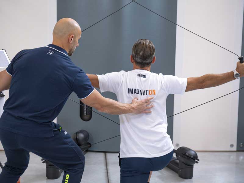 Uomo si allena in palestra concentrandosi su collo, schiena e spalle, seguito da un personal trainer esperto che lo guida nell’esecuzione.