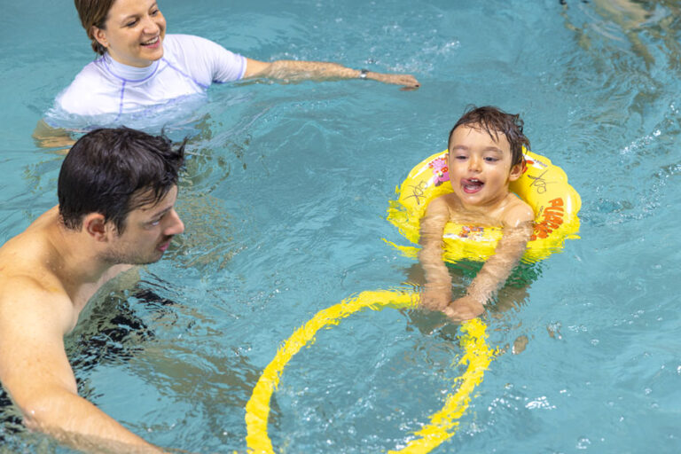 bambino in piscina a padova show club impara a nuotare. sta usando il salvagente ed è affiancato dal papà e dalla istruttrice