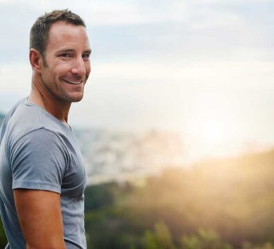Uomo all’aperto in un parco al tramonto, visto di spalle ma con il volto girato verso la fotocamera, sorride ed è vestito in abbigliamento sportivo, mostrando motivazione e prontezza per l’allenamento.