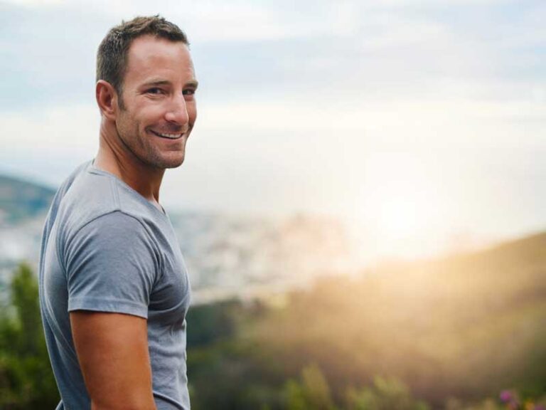 Uomo all’aperto in un parco al tramonto, visto di spalle ma con il volto girato verso la fotocamera, sorride ed è vestito in abbigliamento sportivo, mostrando motivazione e prontezza per l’allenamento.