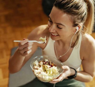 Donna vestita sportiva indossa cuffiette e mangia da una ciotola cibo sano, tra cui verdura e frutta secca.