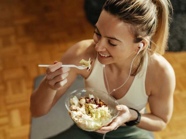 Donna vestita sportiva indossa cuffiette e mangia da una ciotola cibo sano, tra cui verdura e frutta secca.