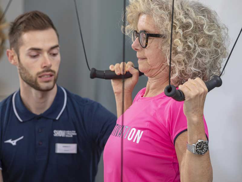 Donna in palestra esegue un allenamento di forza al Kinesis per contrastare l’artrosi, affiancata dal personal trainer che la guida.